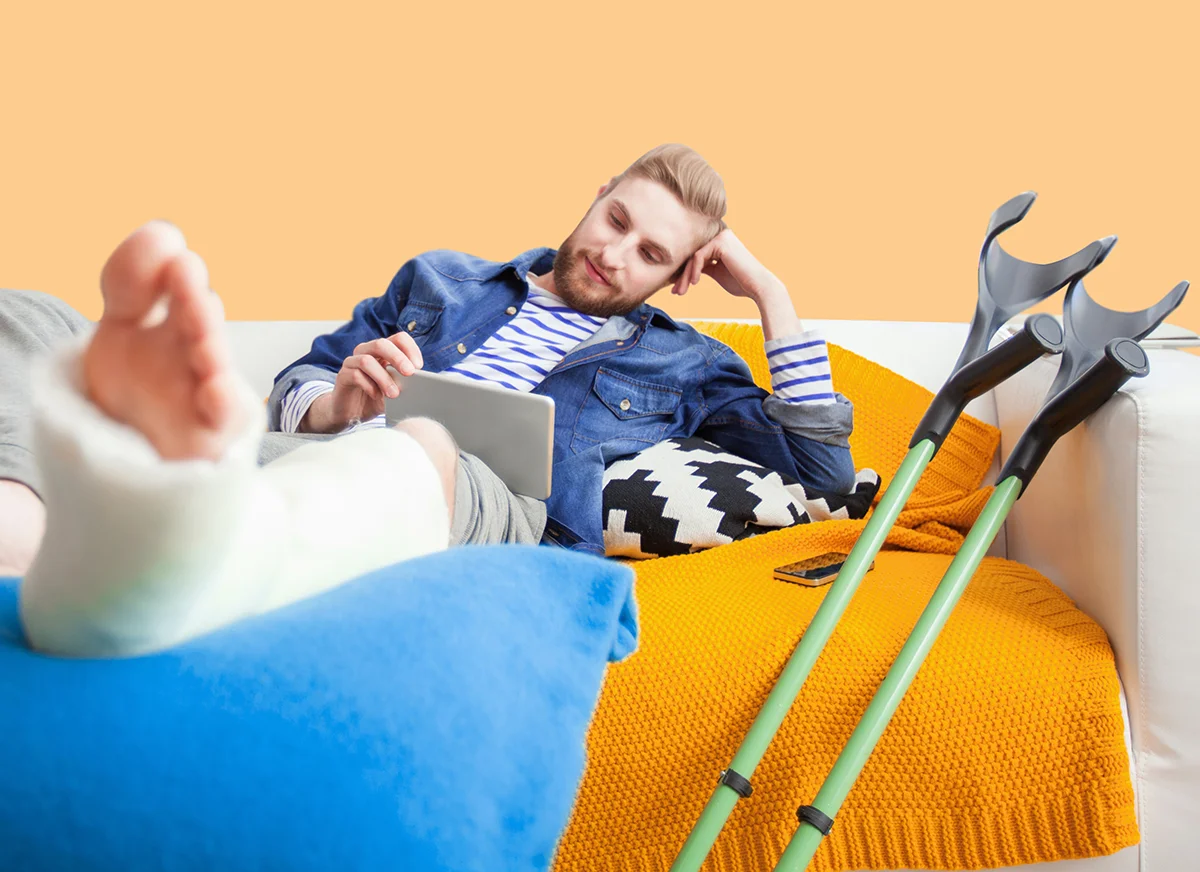 Photographie d'un homme avec une jambe dans le plâtre sur le canapé avec une tablette